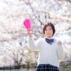 白いカーディガンとチェック柄のスカートを着た学生が、桜に囲まれた屋外に立って、ピンク色のメガホンを持ち、笑顔で拳を握っている。.