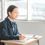 教室で制服を着た女学生が机に座りノートを取っている様子。明るい窓から自然光が差し込む。