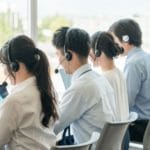 4人が机に並んで座り、それぞれヘッドセットを装着してノートパソコンで作業している。大きな窓から外の景色が見えるモダンなオフィスにいるようだ。.