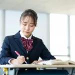 紺色のブレザーと赤いチェック柄のリボンをつけた制服を着た生徒が机に座り、赤いペンで何かを書いています。彼女の横には開いた本が置いてあります。ぼやけた背景は教室の明るい雰囲気を醸し出しています。.