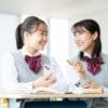 鎌倉市の塾で、制服姿の女子生徒2人が机に座り、スマートフォンを手に笑顔で会話を交わしている。目の前には本が開かれ、楽しそうに会話をしている。.