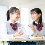 鎌倉市の塾で、制服姿の女子生徒2人が机に座り、スマートフォンを手に笑顔で会話を交わしている。目の前には本が開かれ、楽しそうに会話をしている。.