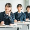 鎌倉市の明るい塾の教室で、制服を着た3人の生徒が机に座っている。前にいる女子生徒は笑顔でノートに書き込み、後ろにいる2人のクラスメートは熱心に授業を聞いている。