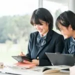 明るい教室で、制服姿の2人の生徒が、本やノートのある机に座りながら、笑顔で一緒にタブレットを見ている。.