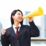 背広姿の女性が微笑みながら黄色いメガホンを口に当て、話しかけたり歓声を上げたりしているように見える。ぼやけた背景に近代的な高層ビルが建ち並ぶ屋外に立っている。.