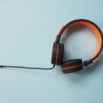 水色の背景に黒とオレンジの有線オーバーイヤーヘッドホンが配置され、ケーブルが画像の左側に伸びている。.