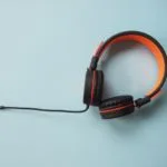 水色の背景に黒とオレンジの有線オーバーイヤーヘッドホンが配置され、ケーブルが画像の左側に伸びている。.