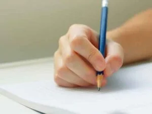 鎌倉市の塾で勉強中の人物と思われる、青い鉛筆を持ち、白い紙に文字を書いている人物の手のクローズアップ。背景はぼかされている。.