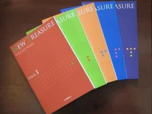 鎌倉市内の多くの塾では対策が不十分な私立中高一貫校で使われている「ニュー・トレジャー・イングリッシュ・シリーズ」のカラフルな教科書5冊が、木の板の上に扇状に広げられている。表紙にはステージ1からステージ5まで、オレンジ、赤、緑、青、紺色のラベルが貼られている。.