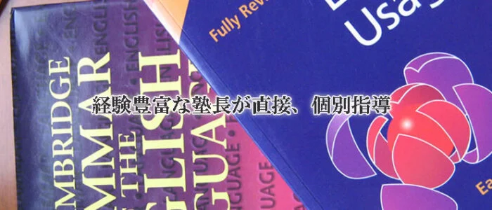 鎌倉市の塾の教室の机の上に、ケンブリッジ英語の教科書 2 冊が重ねて置かれているクローズアップ。近くには鉛筆があり、教科書の上には日本語のテキストが重ねて表示されている。