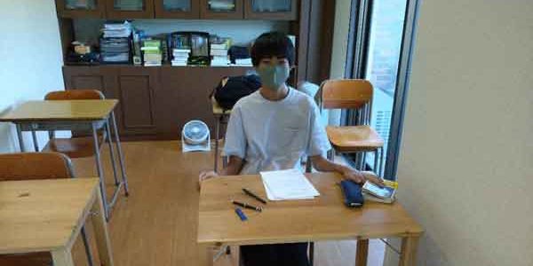 マスクを着けた学生が勉強中の教室。机、ノート、ペンが見える静かな学習環境。